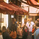Pessoas frequentam restaurantes ao ar livre em uma rua movimentada de Sevilha, enquanto músicos se apresentam.