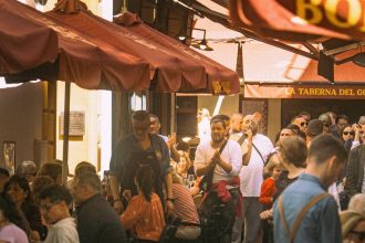 Pessoas frequentam restaurantes ao ar livre em uma rua movimentada de Sevilha, enquanto músicos se apresentam.