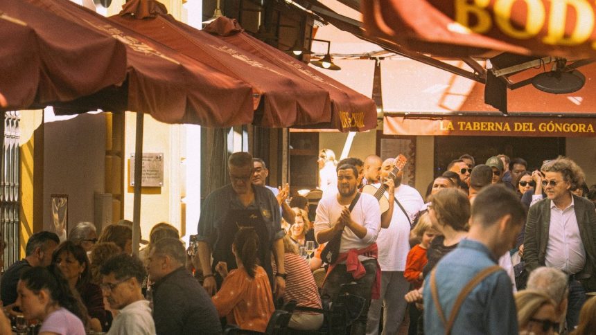 Pessoas frequentam restaurantes ao ar livre em uma rua movimentada de Sevilha, enquanto músicos se apresentam.