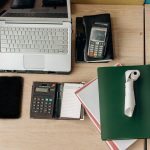 Mesa de escritório com laptop, calculadora, terminal de pagamento e documentos relacionados a atividades financeiras.