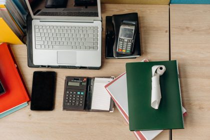 Mesa de escritório com laptop, calculadora, terminal de pagamento e documentos relacionados a atividades financeiras.