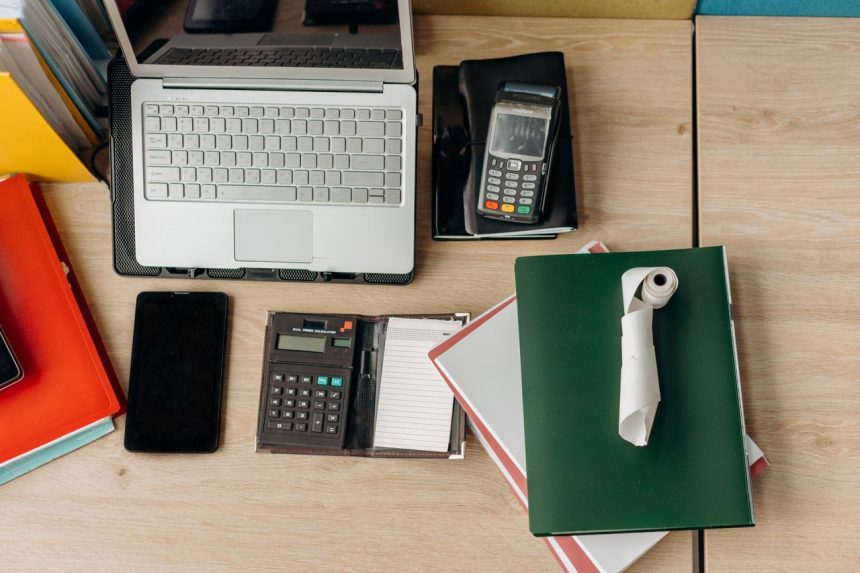 Mesa de escritório com laptop, calculadora, terminal de pagamento e documentos relacionados a atividades financeiras.