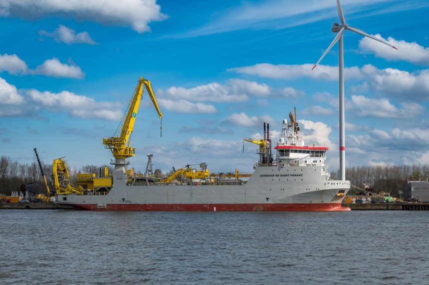 Embarcação industrial atracada em um porto, com uma turbina eólica visível ao fundo.
