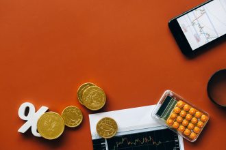Moedas, calculadora e gráficos sobre uma superfície vermelha, representando análise financeira e indicadores econômicos.